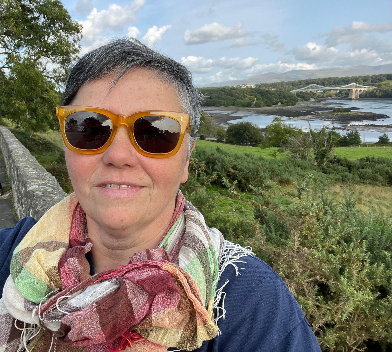 Karen Houghton on Anglesea. A woman wearing yellow sunglasses and a multicolored scarf, taking a selfie outdoors with a river, green landscape, and bridge in the background.