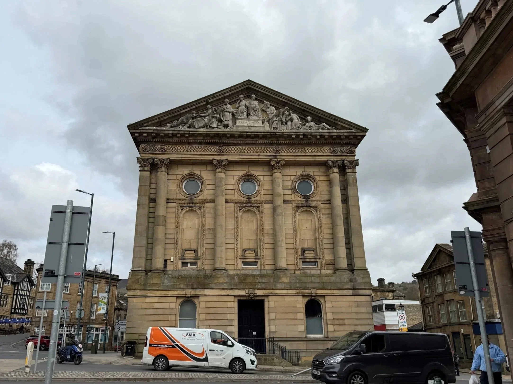 Toddmorden Town Hall. A large historic stone building with classical architecture, featuring a detailed frieze at the top and columns, situated on a busy street with parked cars, vans, and pedestrians, under cloudy skies.