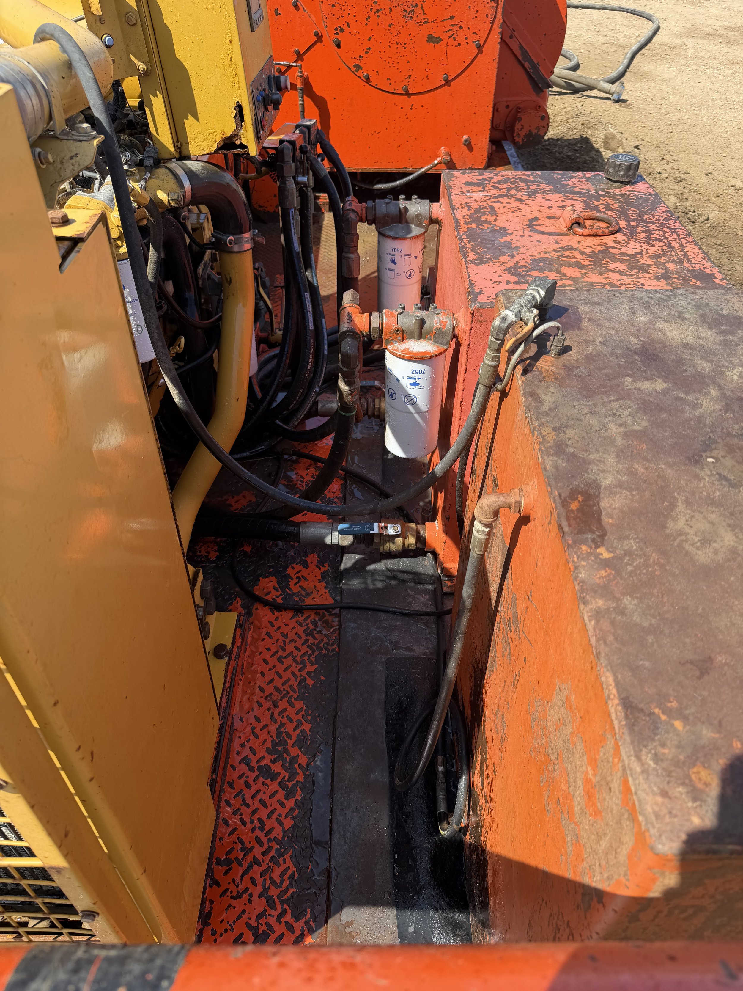 Close-up view of mechanical components, filters, and hydraulic hoses on an industrial orange and yellow machinery, exposed to outdoor environment with dirt ground.