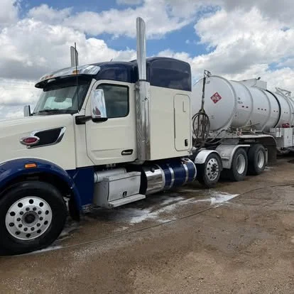 A white and blue semi-truck with a large cylindrical tanker trailer, parked on a muddy ground under cloudy skies.