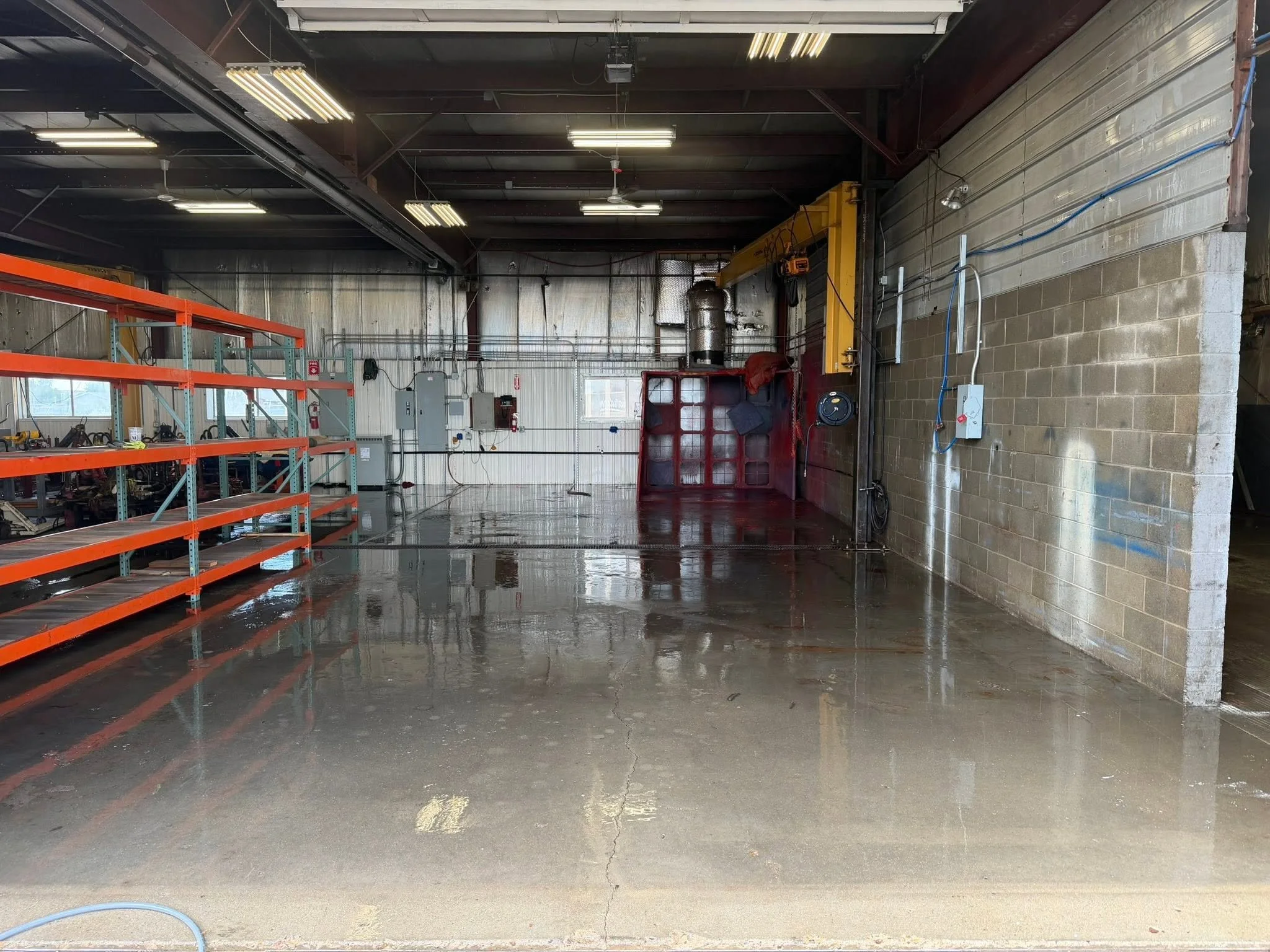 Interior of a warehouse or industrial space with metal shelving on the left, concrete and brick walls, fluorescent ceiling lights, a yellow overhead crane, and wet floors with a drain and some electrical wiring that was pressure washed.