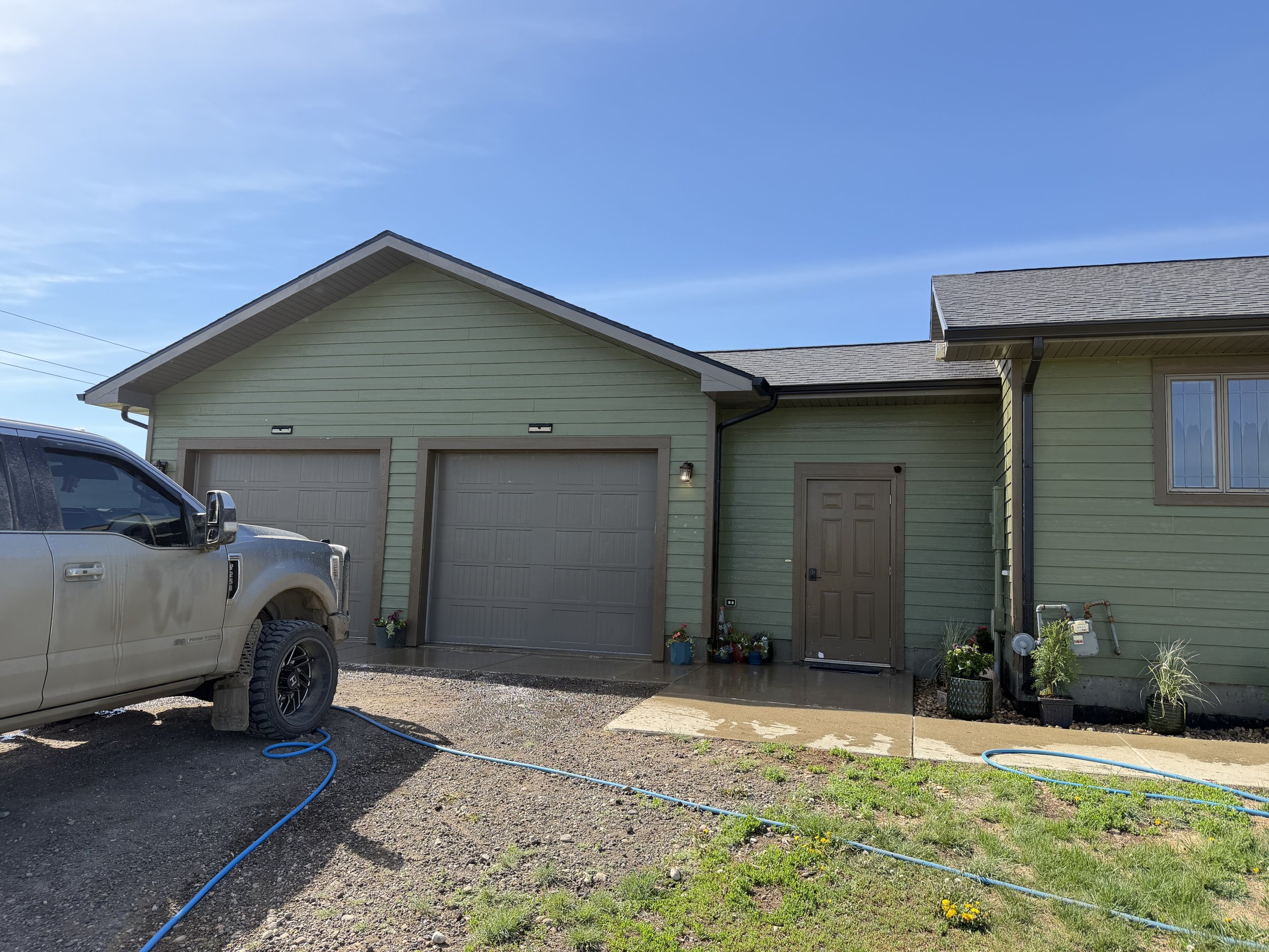 A light green house with a two-car garage and a brown front door, two wall-mounted lights, several potted plants near the entrance, and a window on the right side. A black and white pickup truck is parked in front, with garden hoses on the gravel dri