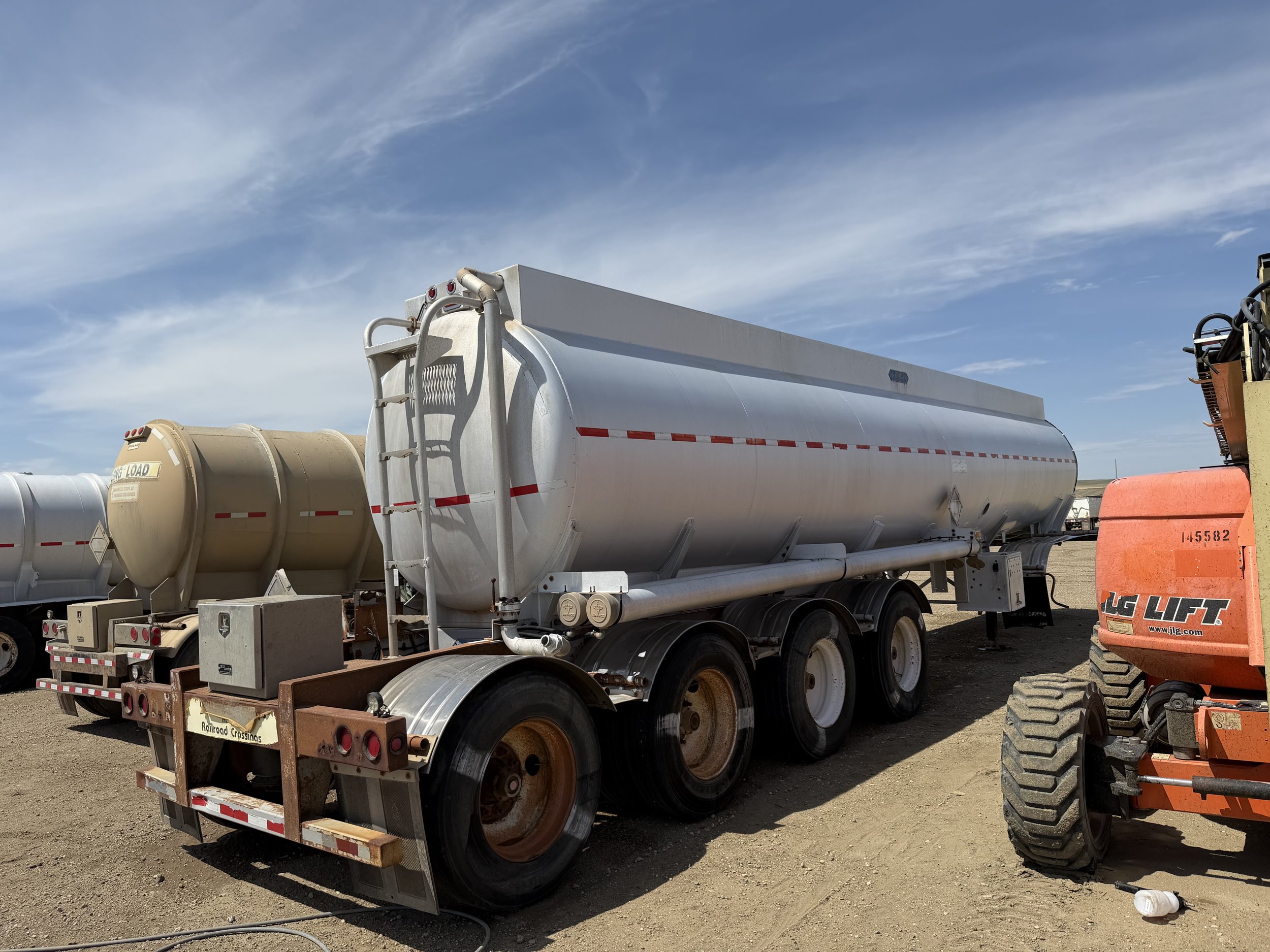 A white tanker truck parked on a dirt lot, with a beige tank in the background and an orange compact excavator to the right, under a partly cloudy sky.