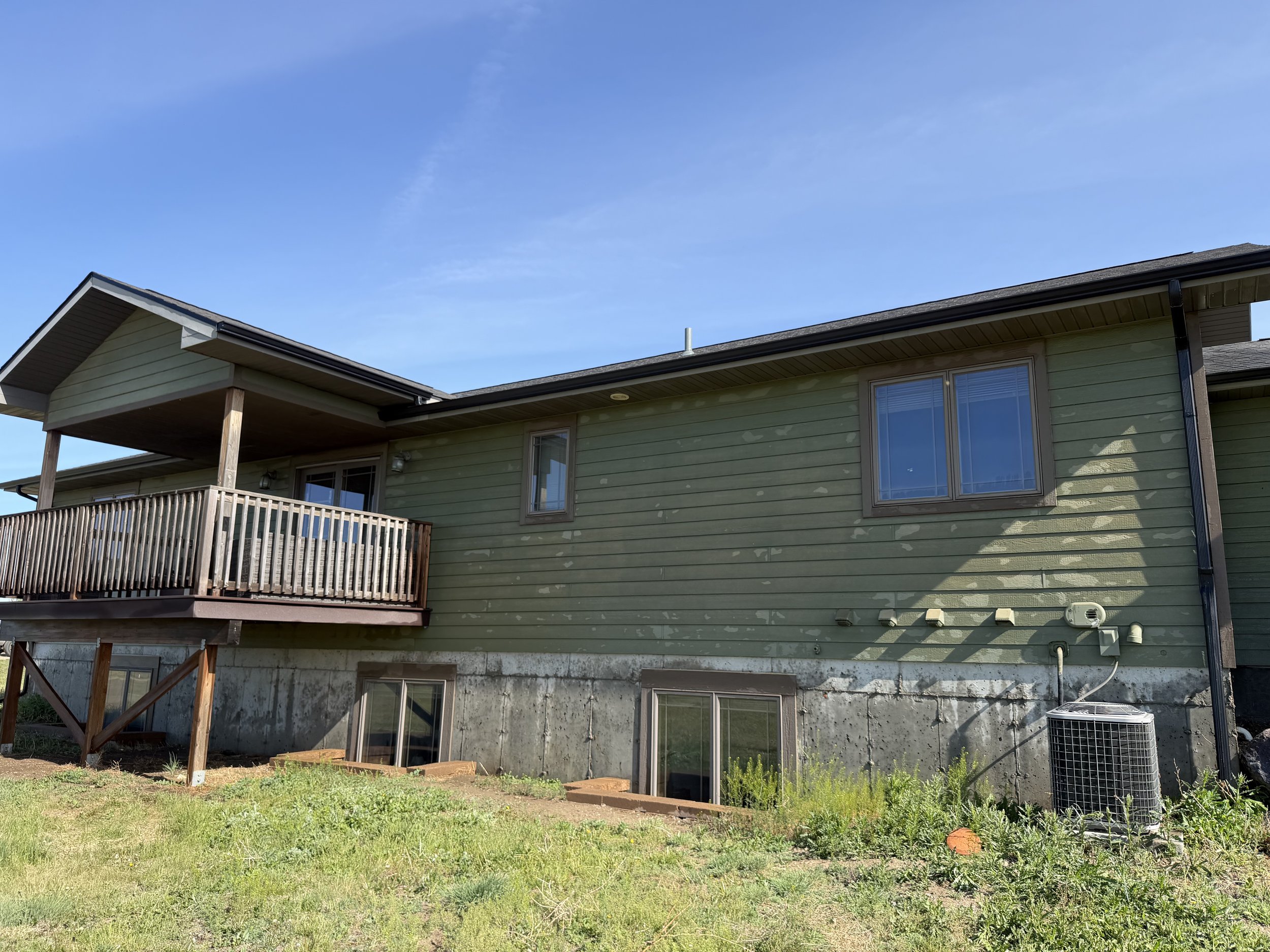 A two-story house with green siding, a wooden balcony, and large windows, with a partly cloudy blue sky overhead.
