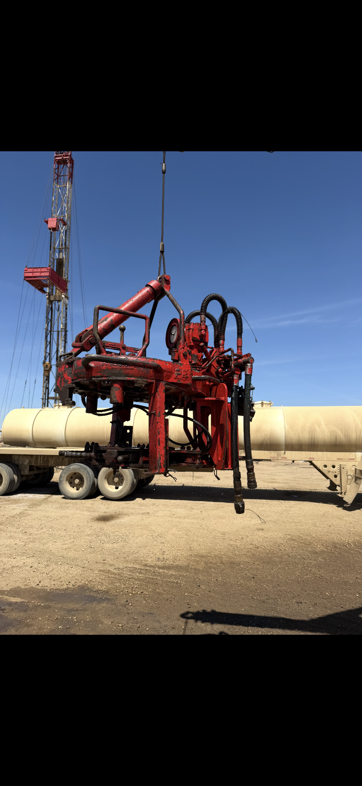 A large red industrial valve or pump system suspended in the air at an oil or gas site, with a drilling rig in the background under a clear blue sky.