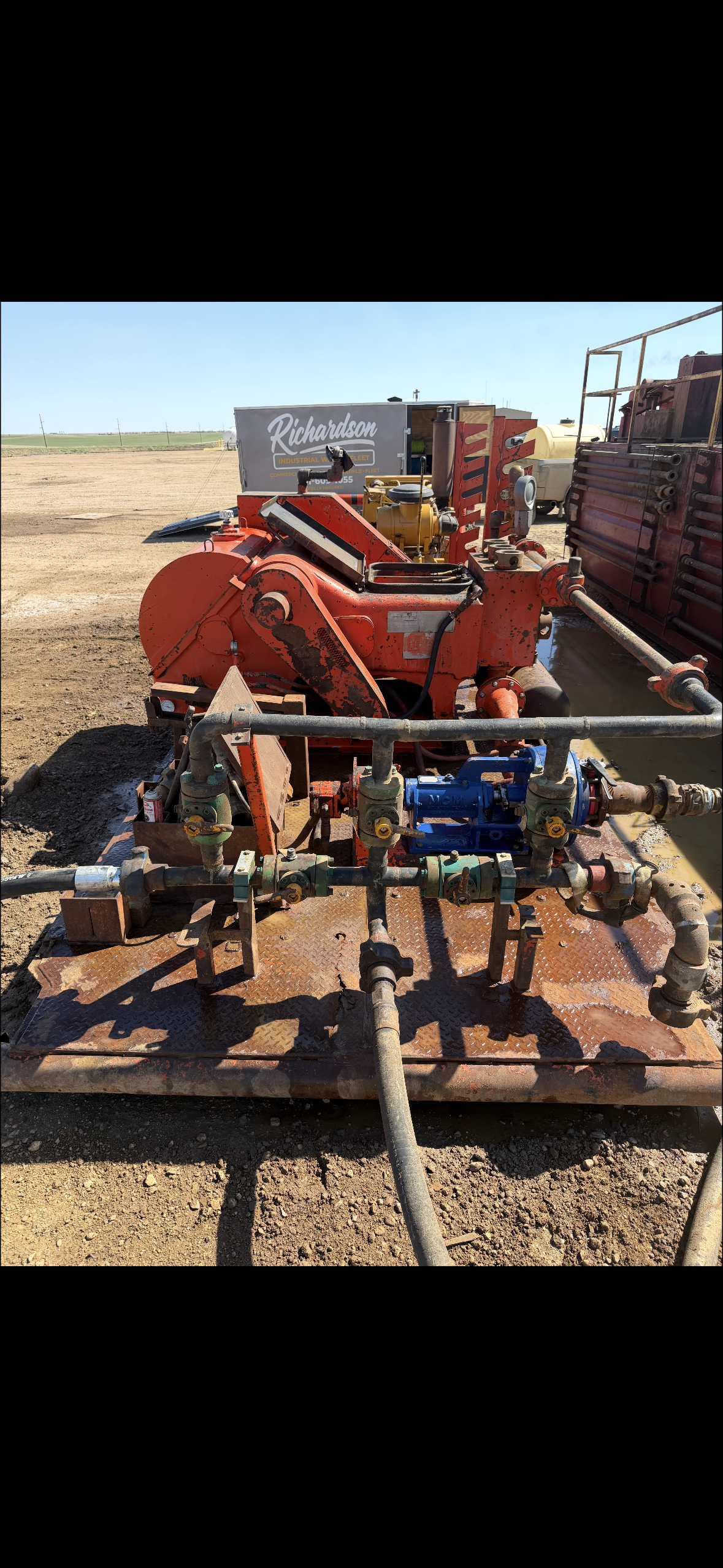 A red industrial machine with pipes and gauges on a metal platform in a dirt field under a clear blue sky.