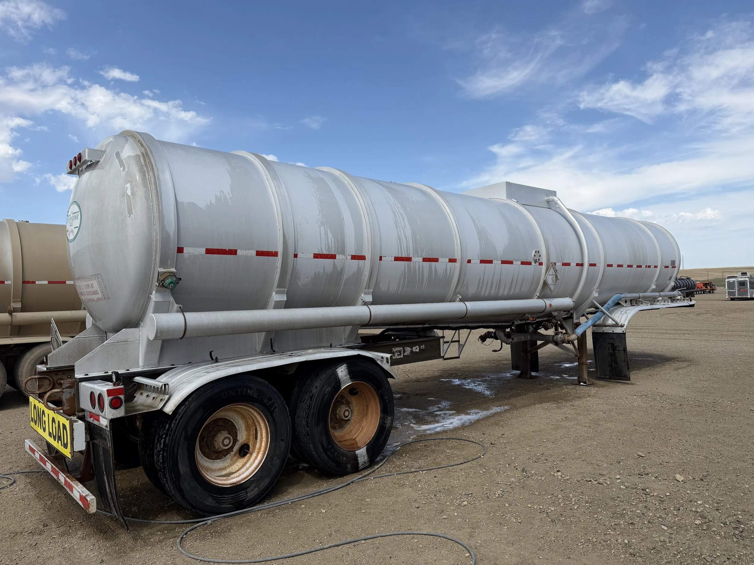 A large white tanker trailer with red and white reflective strips parked on a dirt lot with other trailers and trucks in the background, under a partly cloudy sky.