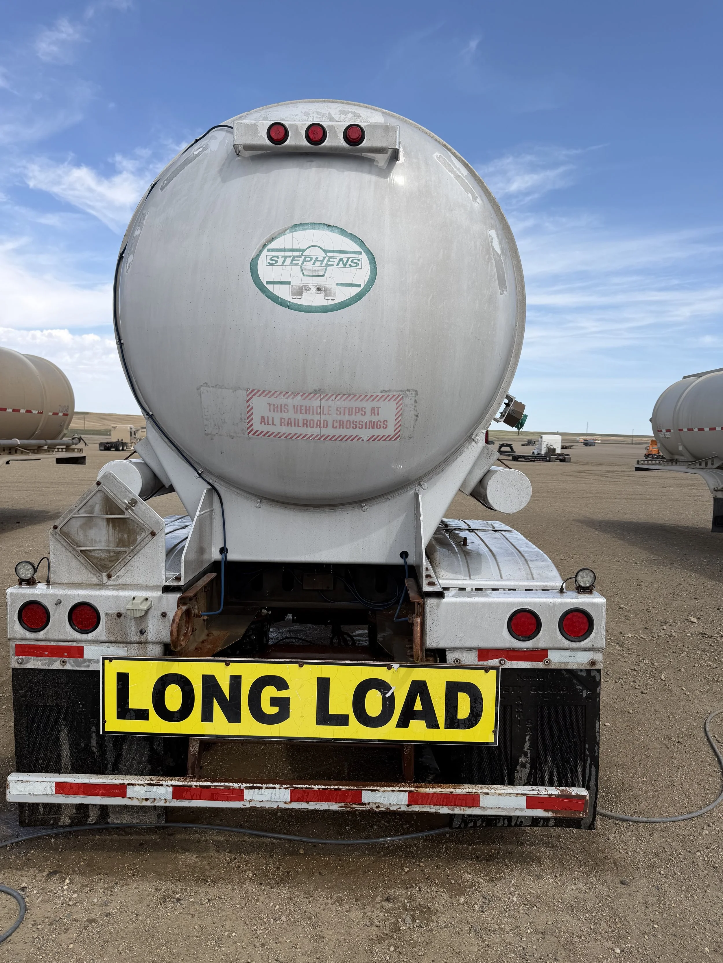 Rear view of a propane or fuel tanker truck with a yellow 'LONG LOAD' sign attached to the back, parked on a dirt lot with other tanker trucks in the background under a partly cloudy sky.