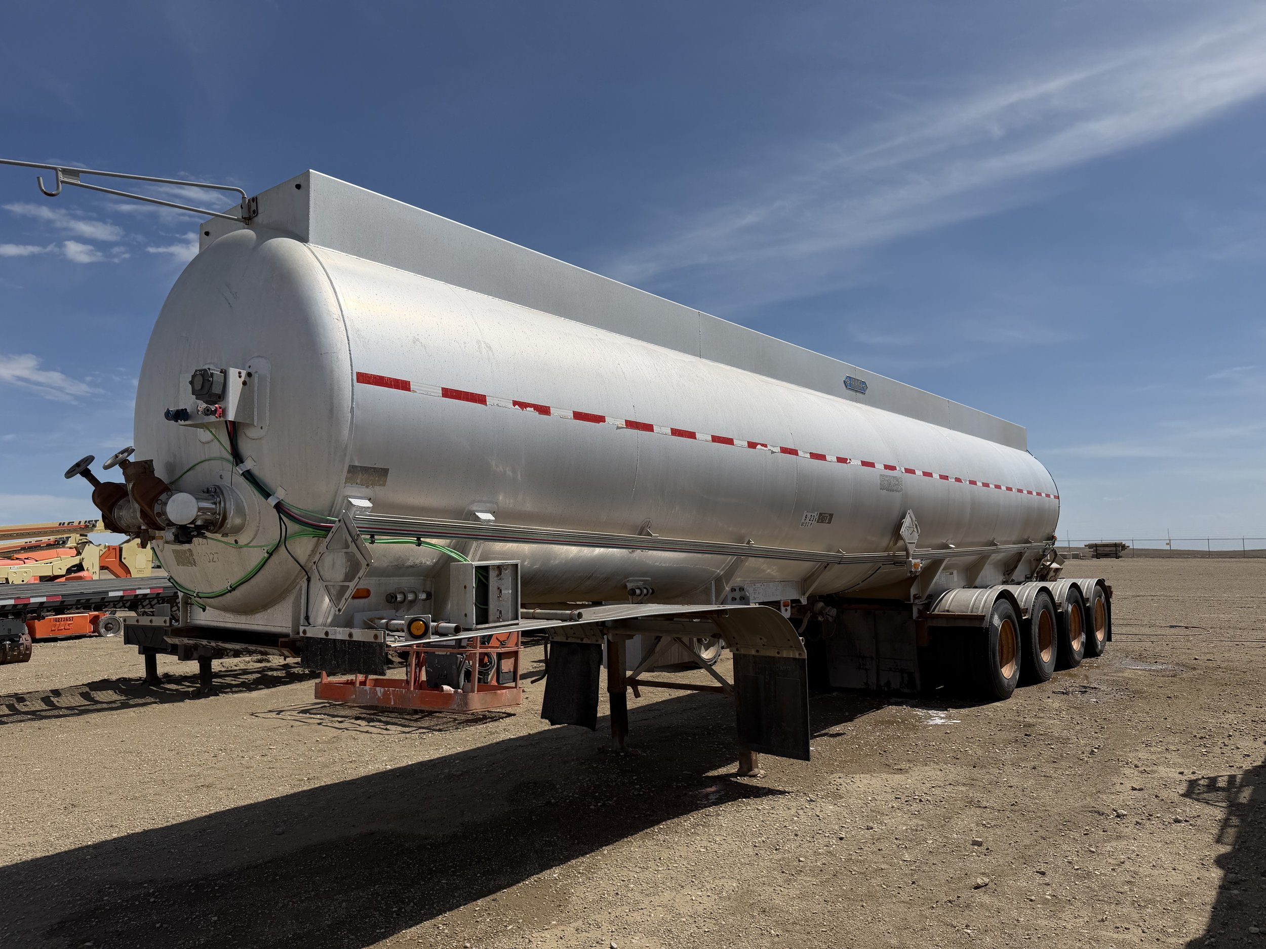 A large white tanker trailer parked on a dirt lot under a blue sky with wispy clouds.