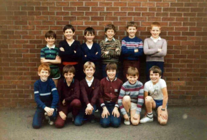 Group of ten school children, five in the back row standing against a brick wall, and five sitting on the floor in front, smiling for a class photo.