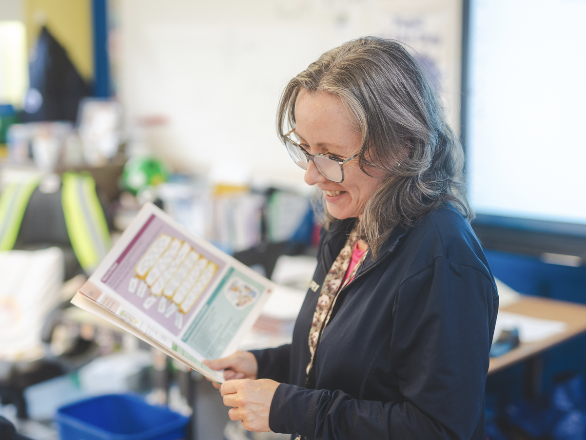 A woman with wavy gray hair and glasses smiling while reading a colorful brochure in an office or classroom setting.