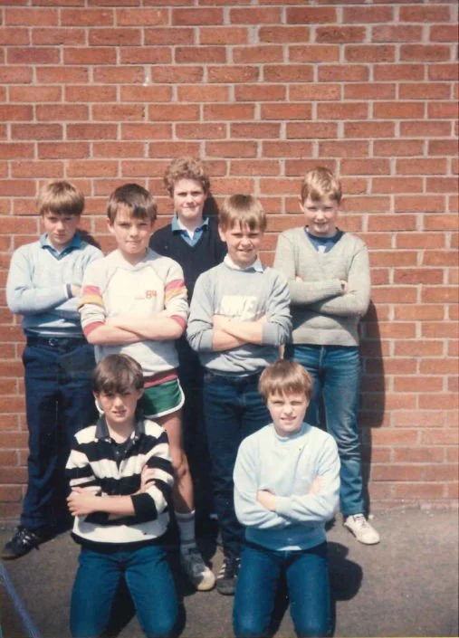 Seven children standing and kneeling in front of a brick wall, posing with crossed arms for a group photo.