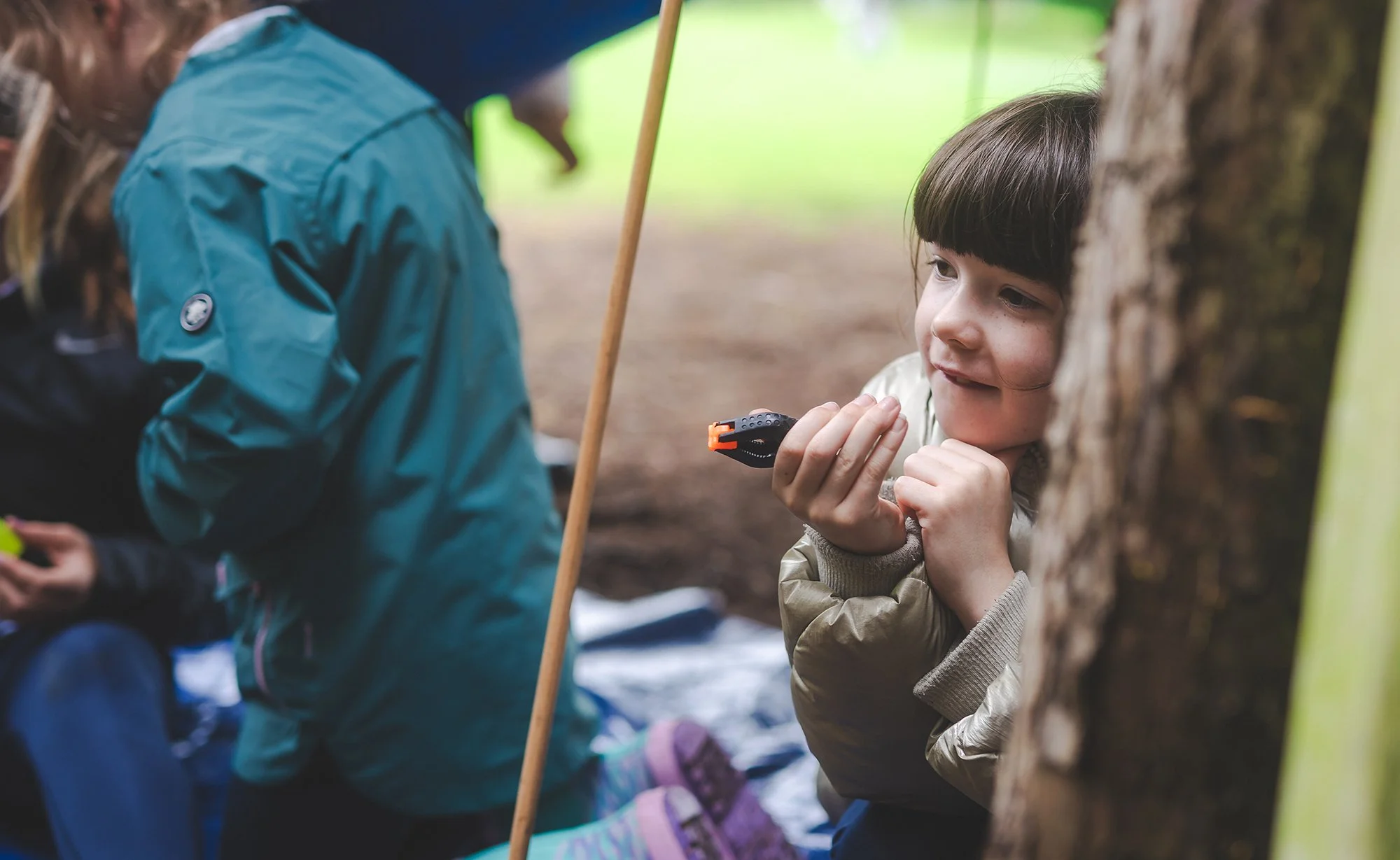 Two children are in a forest, one hiding behind a tree and the other looking at her. The girl is holding a small black device, possibly a toy or a gadget, and wears a beige jacket. The boy is partly obscured, wearing a blue jacket. They appear to be playing or hiding in the woods.