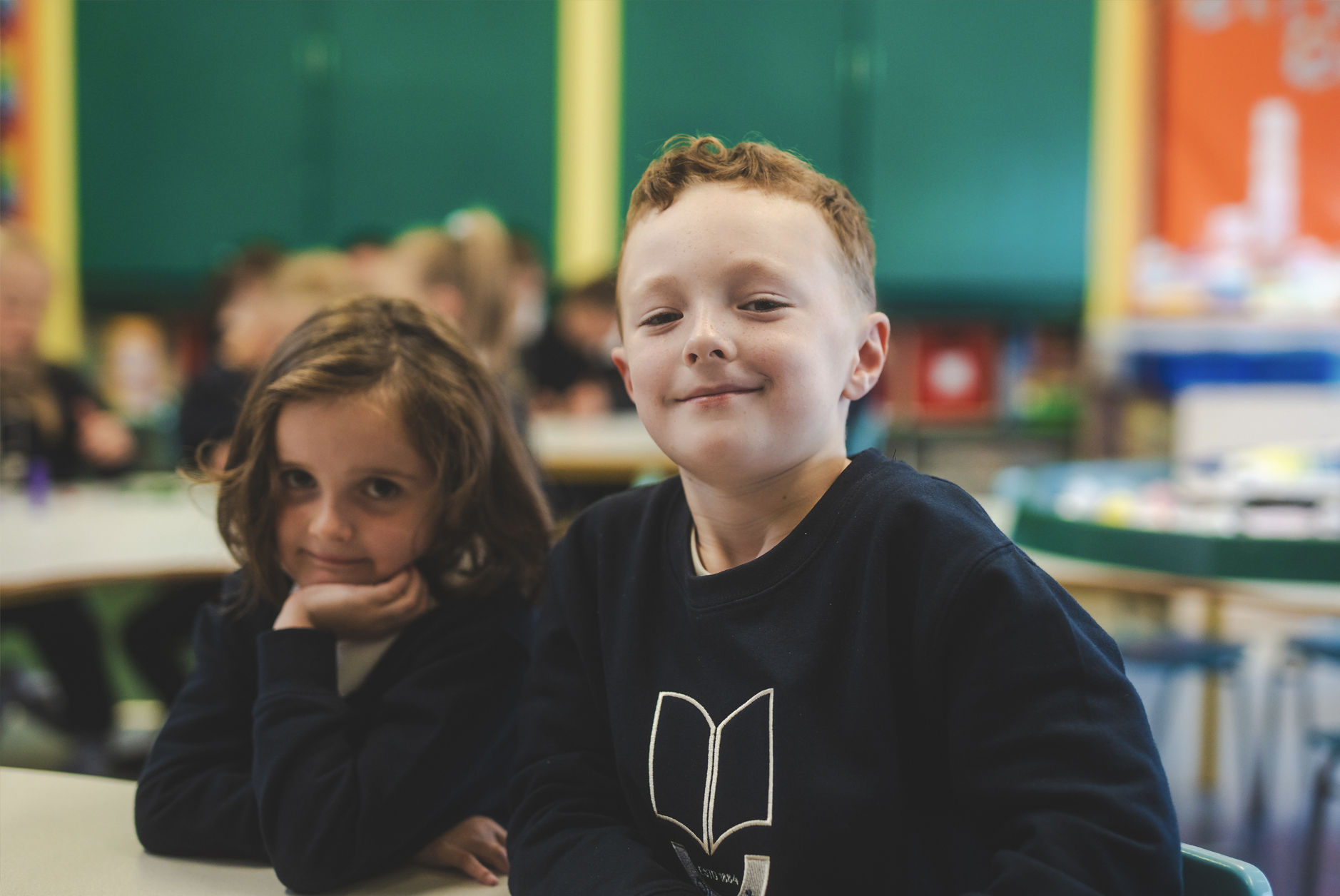 Two children sit at a school desk in a classroom, with a boy smiling and a girl resting her chin on her hand.