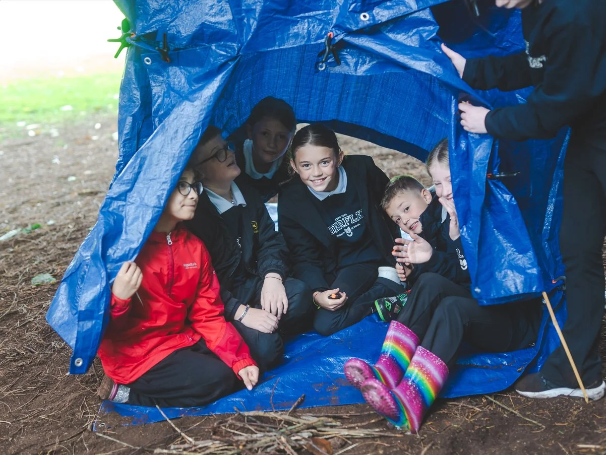 Children sitting inside a blue camping tent outdoors on the ground with dirt and branches, smiling and looking at the camera, dressed in outdoor clothing.