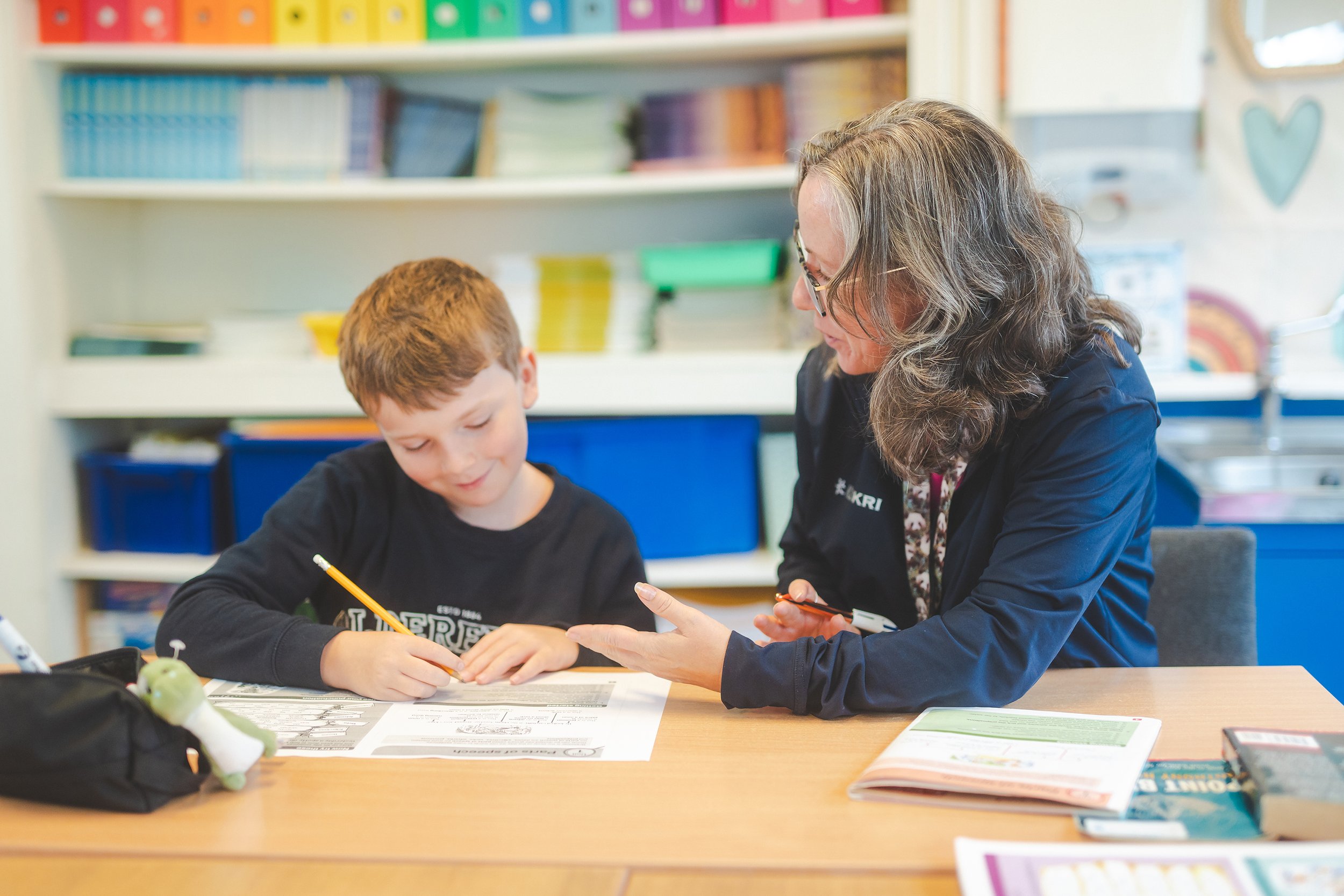 A woman and a young boy sitting at a table in a classroom, engaged in a learning activity. The table has papers, a textbook, and a soft toy. Shelves with colorful storage bins are in the background.