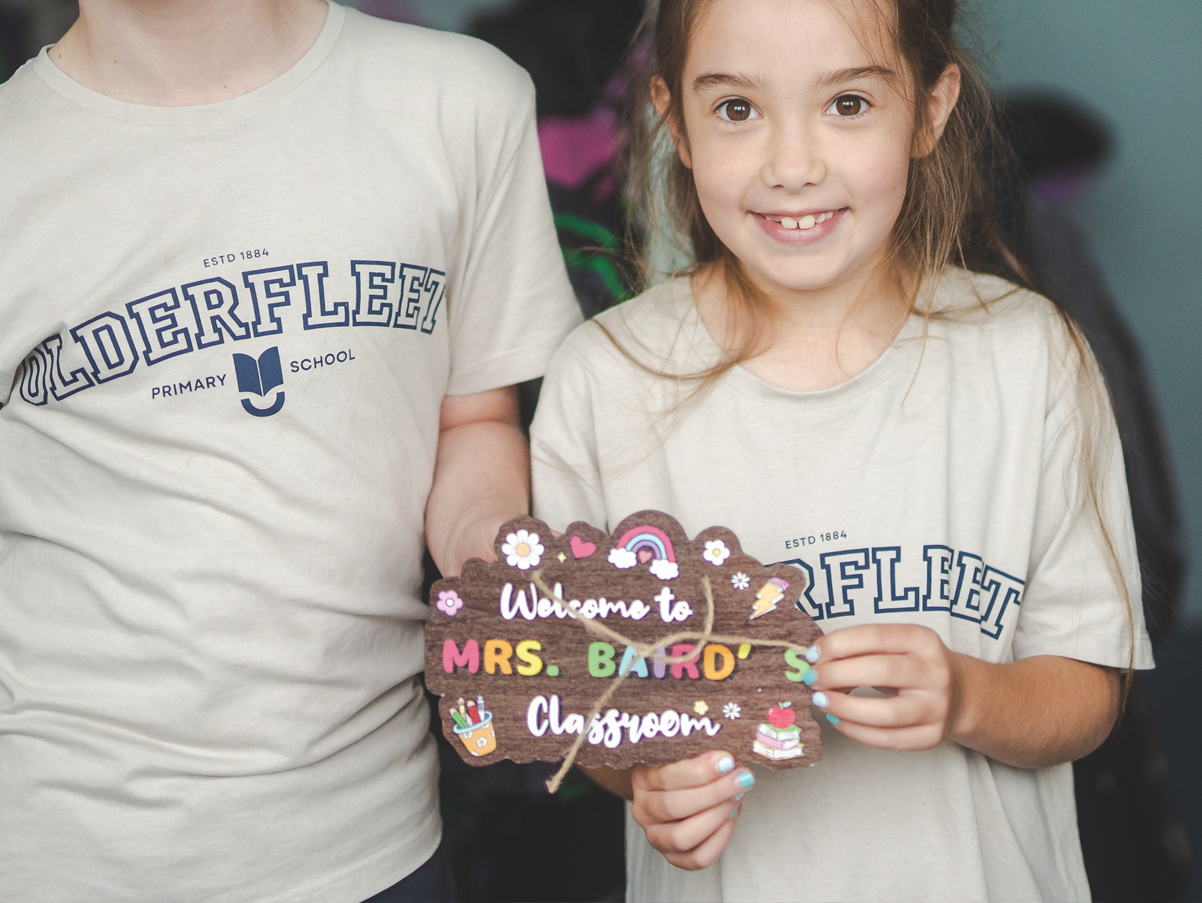 A young girl holding a colorful sign that reads "Welcome to Mrs. Baird's Classroom" with decorative drawings of flowers, a rainbow, a book, a paintbrush, and an apple. She is smiling and wearing a beige T-shirt with the words "Olde Fleet."