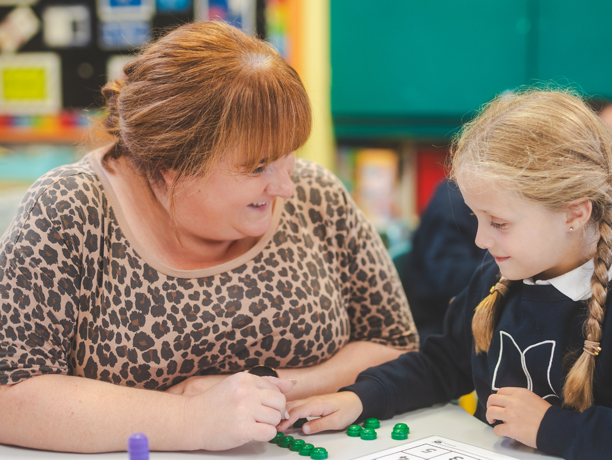 A woman and a schoolgirl playing a game with green tokens at a classroom table, smiling and interacting.