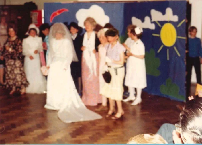 Children performing a play on stage with a painted backdrop of clouds, sun, and trees, while children in costumes look on.