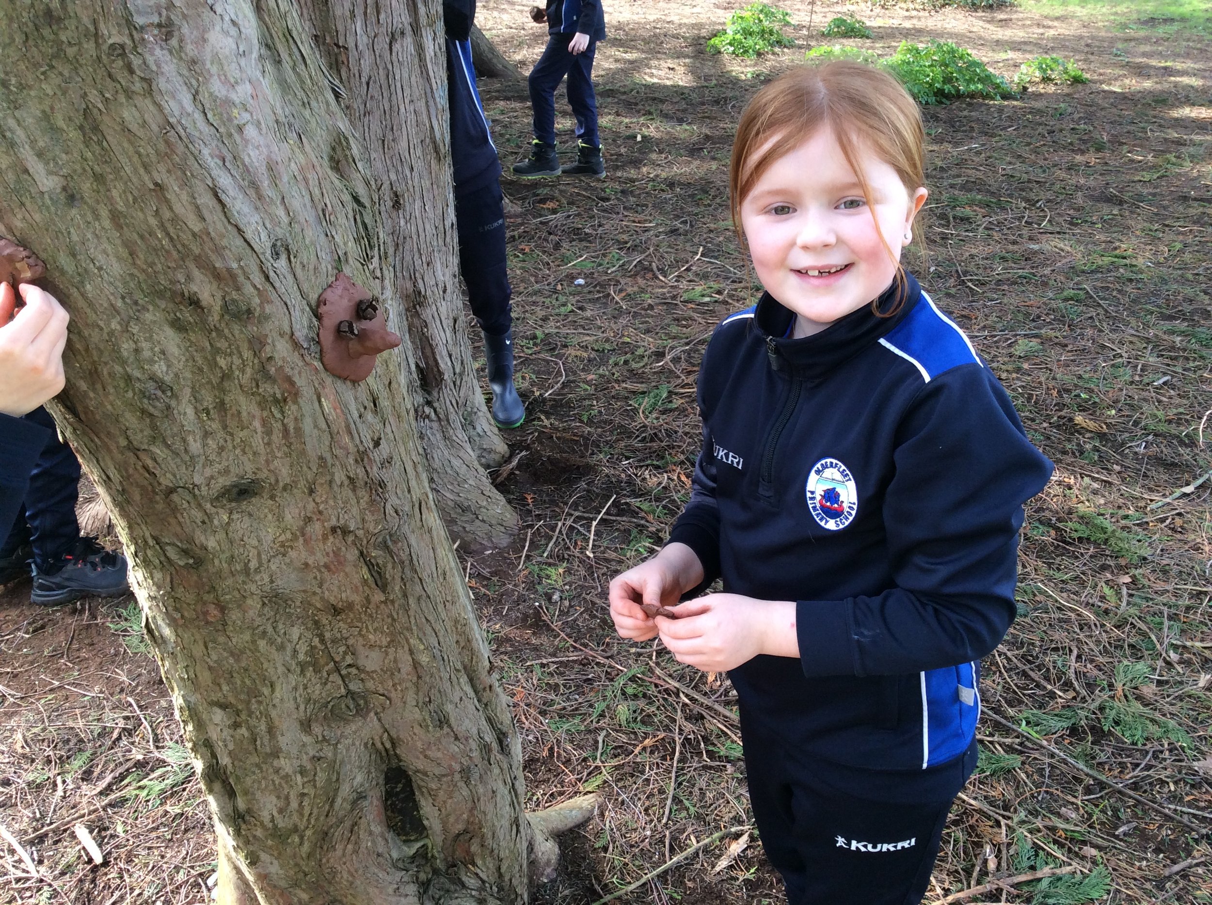 Smiling young girl with red hair and freckles outdoors in a forest, next to a tree with a natural bear-shaped knot in the bark.