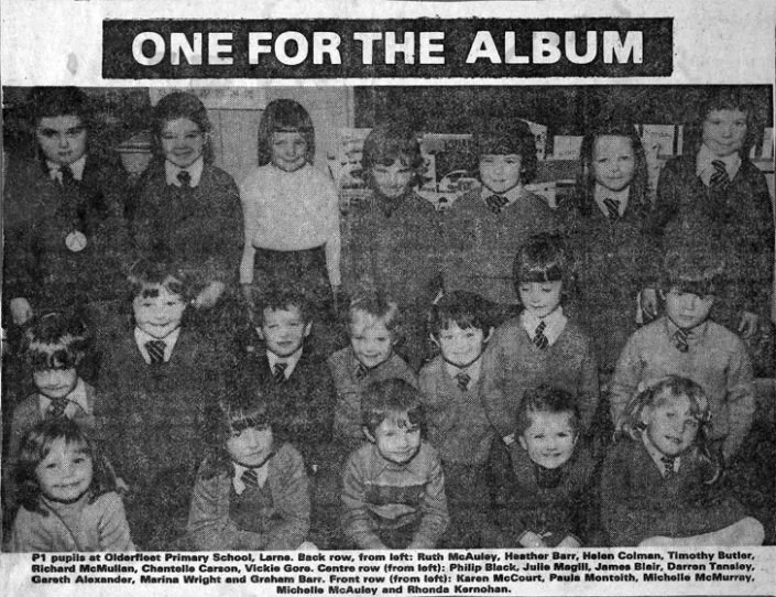 Children in school uniforms posing for a class photo in front of a bookshelf at Olderfleet Primary School, with a sign that reads 'One for the Album.'