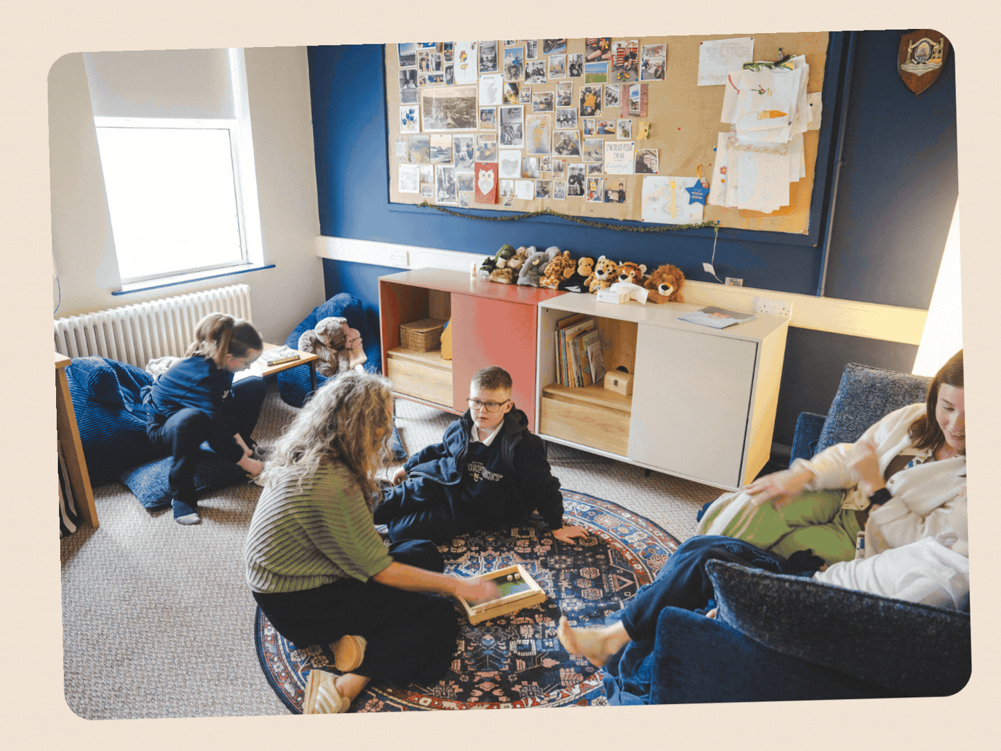 Children and a woman in a classroom setting; some children are sitting on the floor, one girl is on a bean bag, a boy and a woman are on a patterned rug, and another woman is sitting on a chair; storage cabinets with books and stuffed animals are in the background, along with a bulletin board covered with photos and papers.
