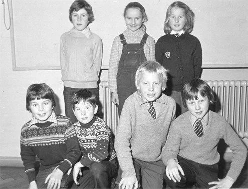 Black and white photo of six children, three sitting in front and three standing behind, in a classroom setting.