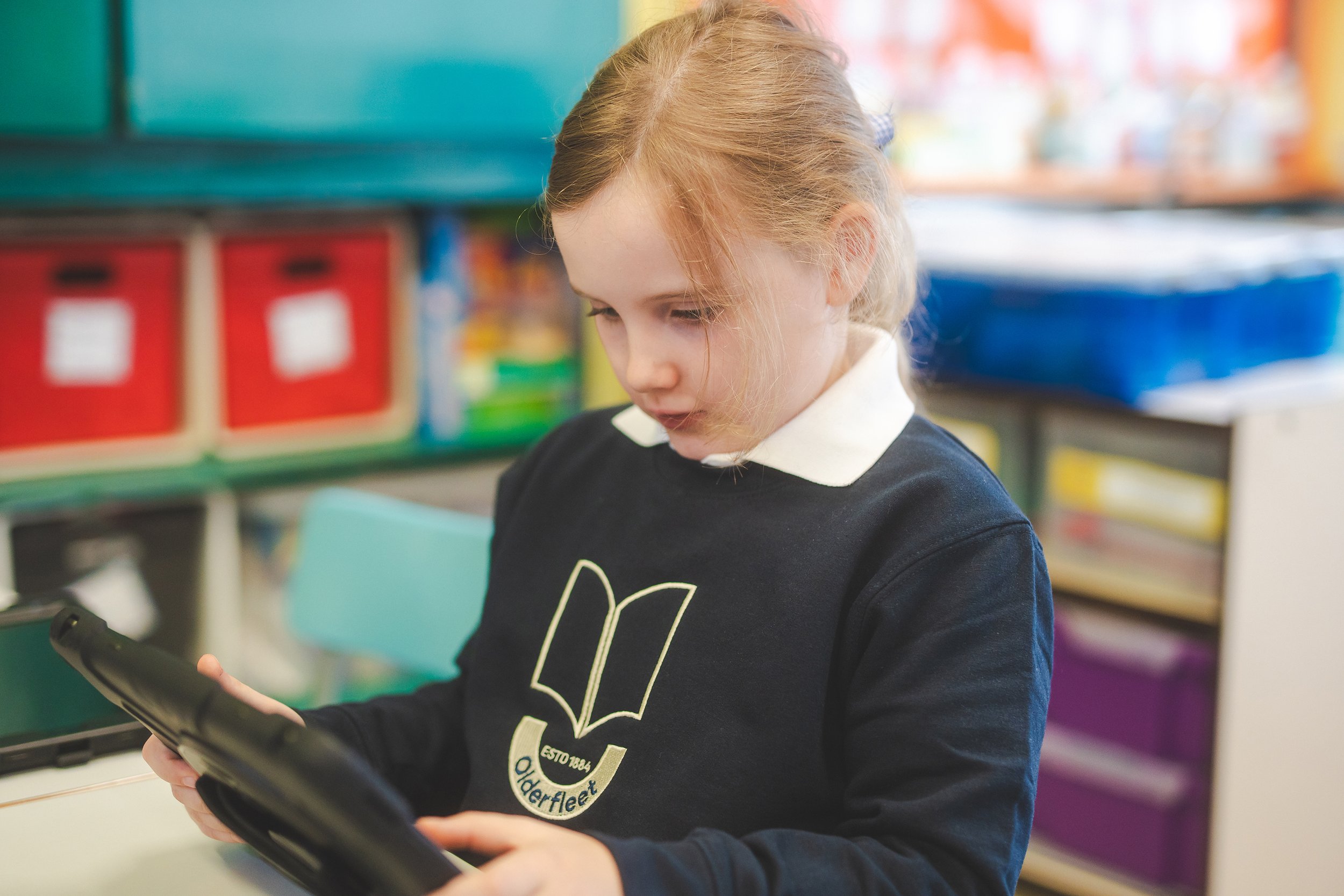 A young girl with blonde hair, wearing a black sweatshirt and a white collared shirt, stands in a classroom looking at a tablet device.