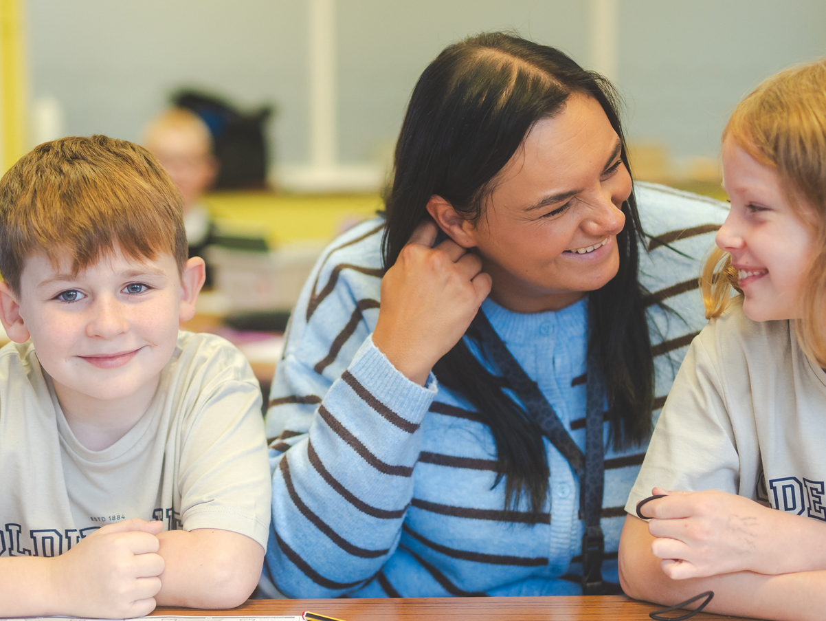 A woman smiling and leaning towards two children at a table, all three engaging in conversation and appearing happy in a classroom setting.