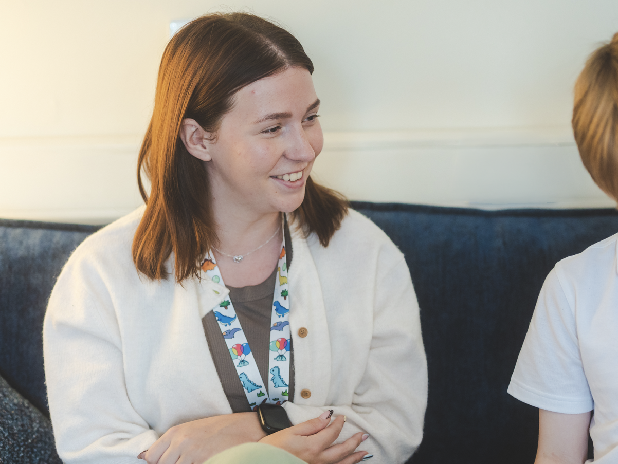 A woman with brown hair smiling during a conversation, wearing a cream cardigan, a lanyard with colorful designs, and a smartwatch, sitting on a blue sofa.