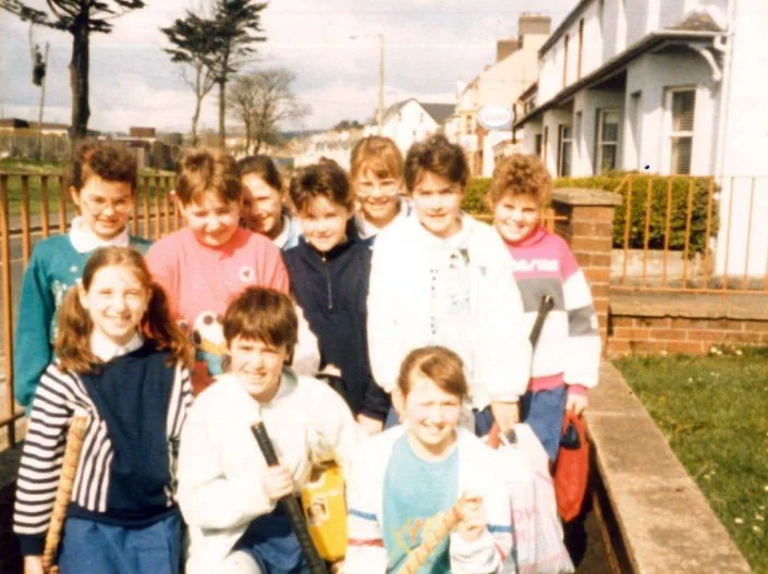 Group of kids standing outside on a sidewalk, smiling for the camera. Some children are holding books or school supplies, and houses are visible in the background.