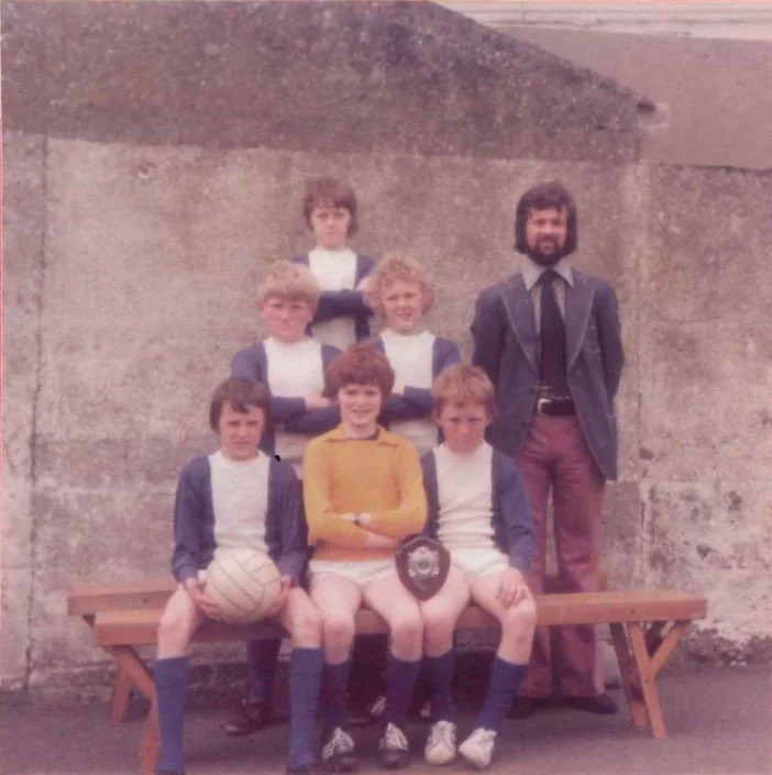 A group of six children and one adult man posing for a photo outdoors in front of a textured wall, with some children sitting on a wooden bench and others standing behind.