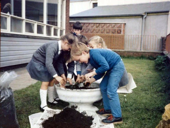 Four children planting a seedling in a large outdoor container, with soil and gardening tools, in a backyard.