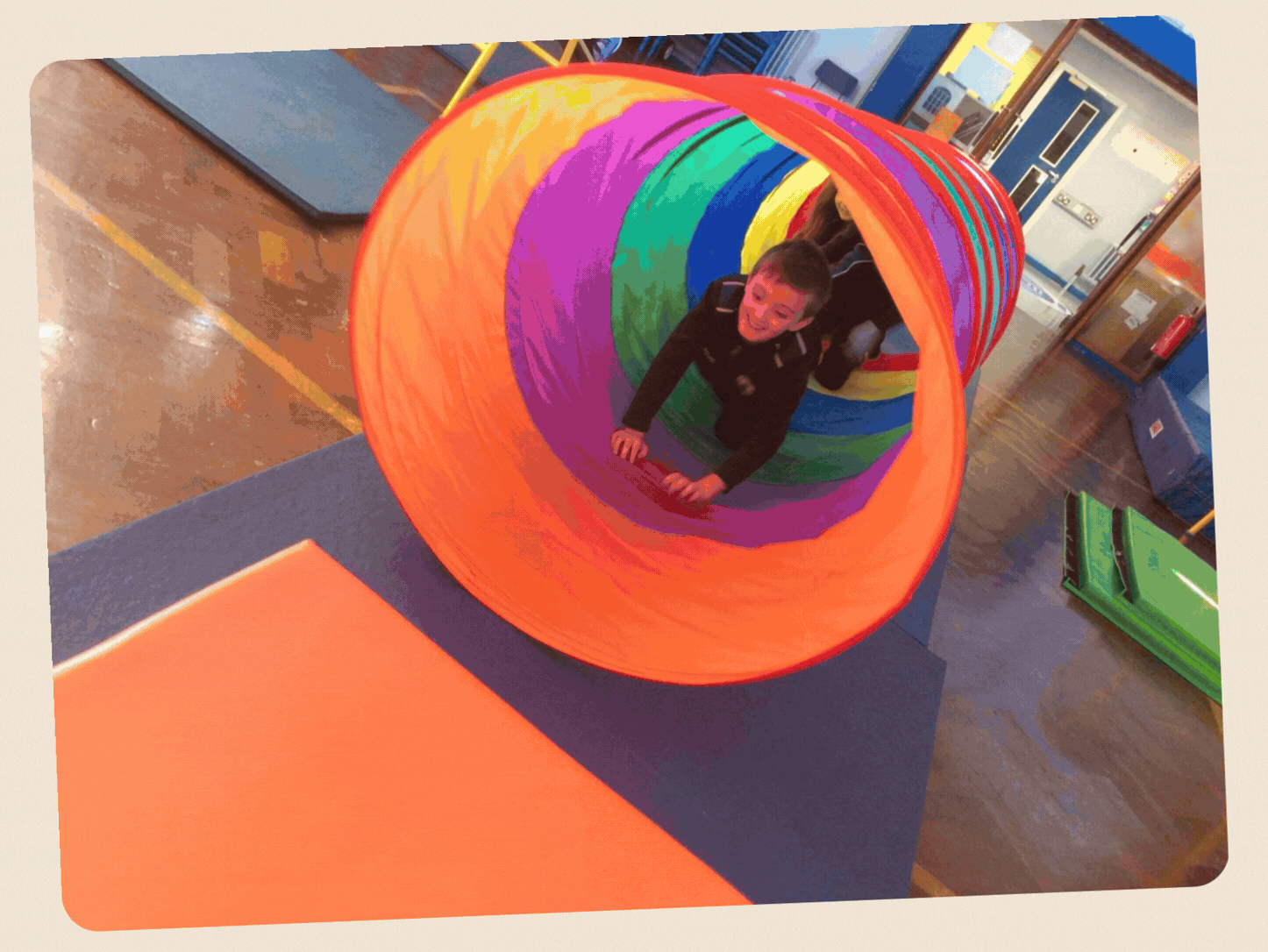 Child crawling through a colorful rainbow-striped fabric tunnel in a classroom or play area.