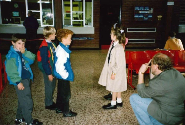 Four children, three boys and one girl, standing in a line, facing an older girl with pigtails, inside a room with red chairs and a carpeted floor, while an adult man sits nearby watching.