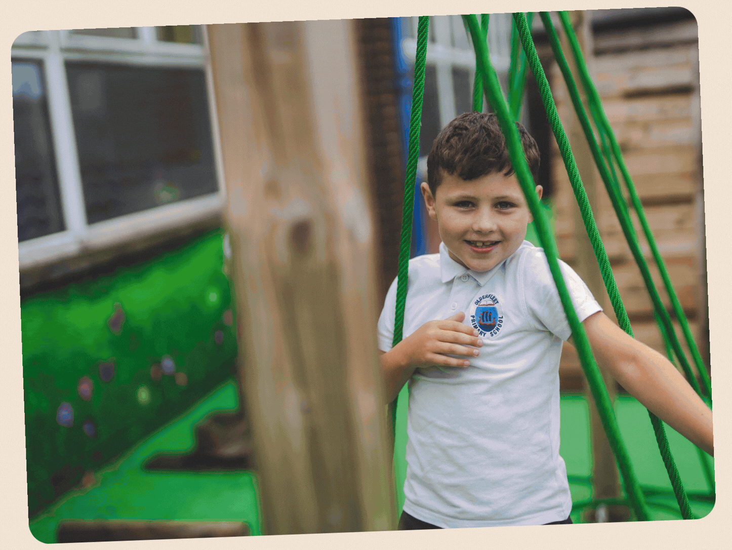 A young boy with short dark hair and a big smile, wearing a white polo shirt with a school logo, standing next to a green playground structure with ropes, outdoors in a backyard with a wooden and brick wall in the background.