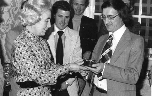 A woman and a man exchanging a small object, with three men watching, in a room with patterned wallpaper and a window in the background.