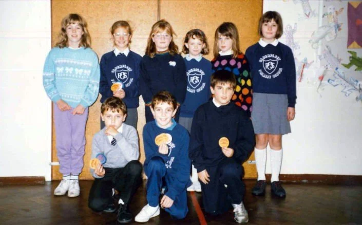 Group of nine children, six girls and three boys, standing and kneeling in a classroom with a brown wall and drawings on the wall behind them. The children are wearing school uniforms or casual clothing.
