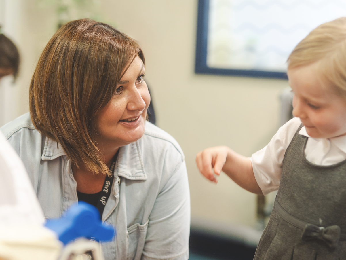 A woman smiling while interacting with a young girl during a conversation in a room with a window.