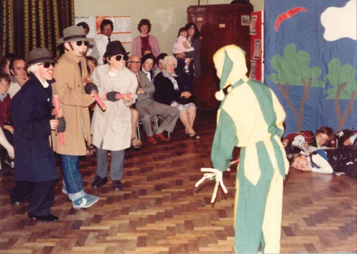 Children dressed in costumes performing a puppet show in front of an audience in a school setting with a colorful painted backdrop.