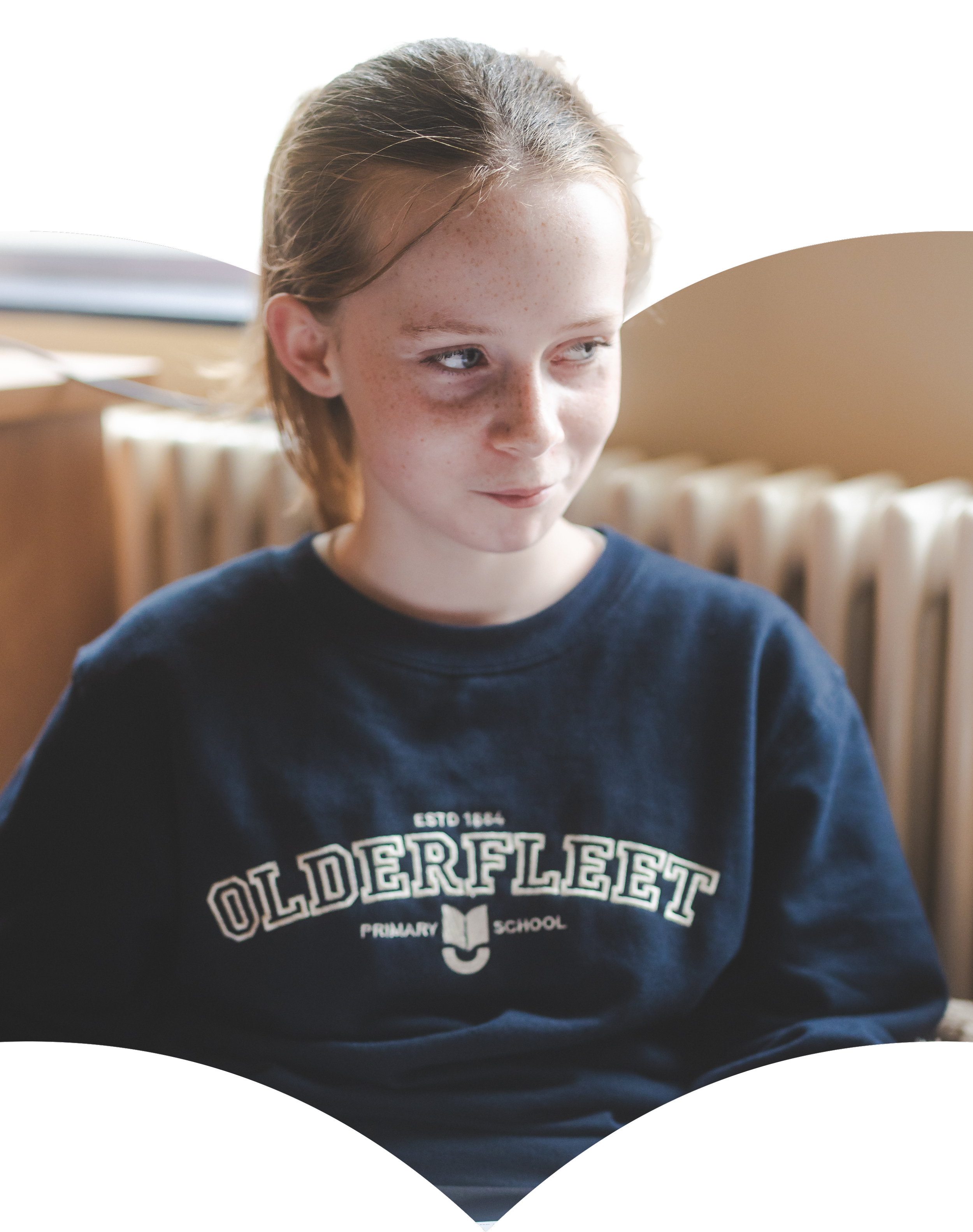Close-up of a young girl with light skin and freckles, wearing a navy sweatshirt with "Oldfield" school logo, in a school setting with blurred background.