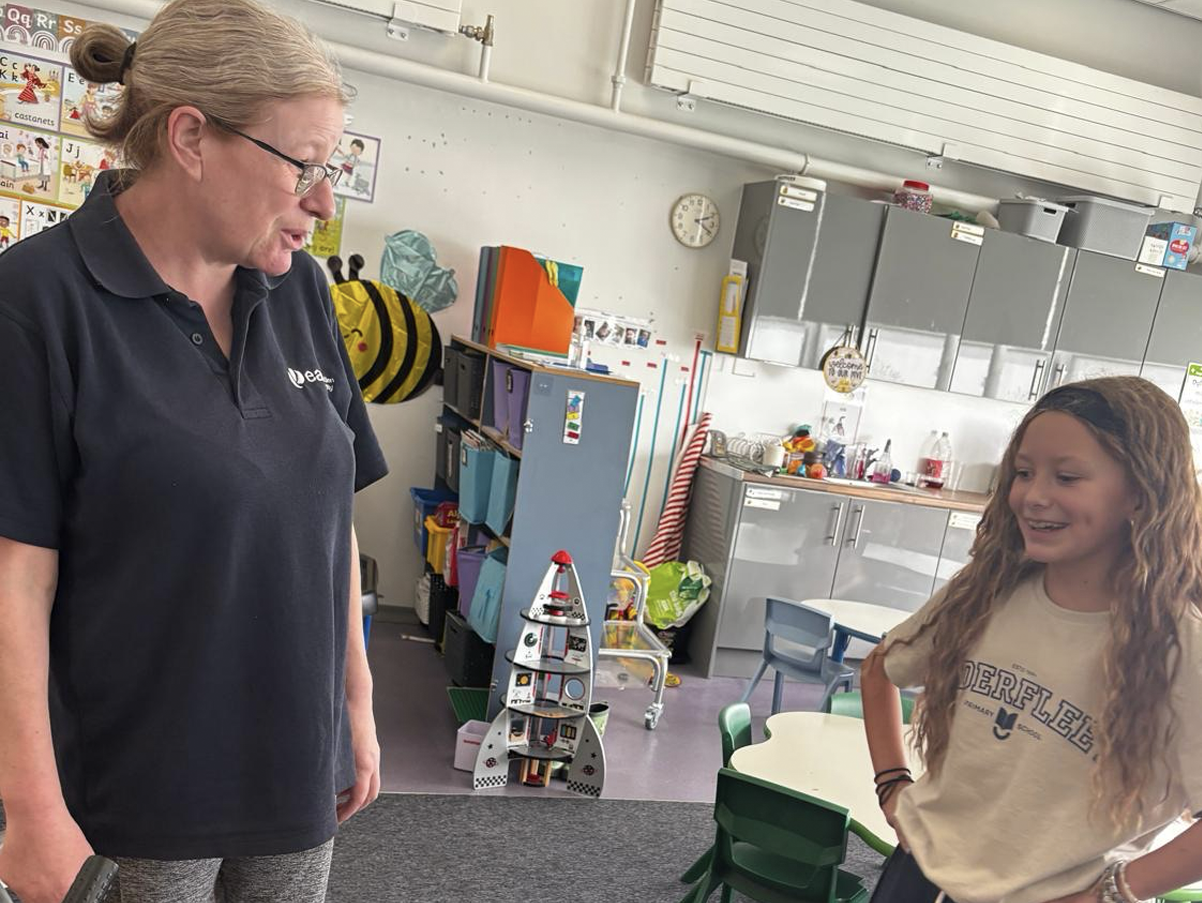 A teacher and a student are having a conversation in a classroom. The teacher, with glasses and a black polo shirt, looks down and speaks while the girl, with long curly hair, smiles and interacts. The classroom background includes storage cabinets, educational posters, and children's toys.