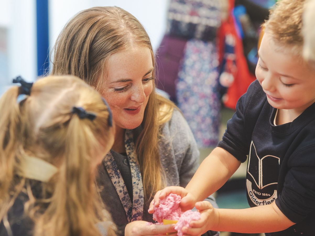 A woman with long blonde hair and freckles is smiling as she assists a young boy with blonde hair and a dark sweatshirt, who is holding a pink fluffy object, possibly playdough or slime, in a classroom or activity setting. Two young girls with blonde hair tied in pigtails are observing.