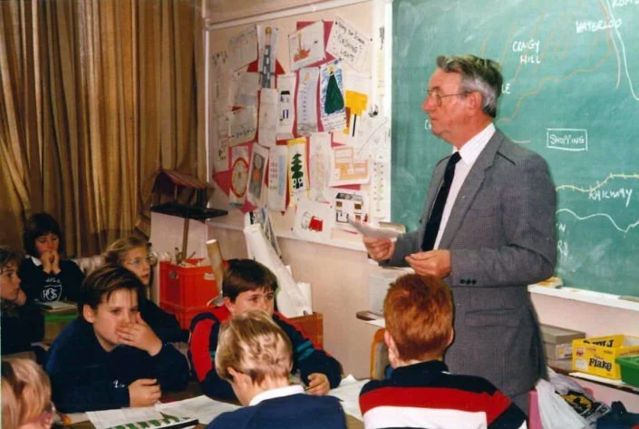 A teacher in a gray suit standing in front of a classroom blackboard, holding papers while students sit at desks and listen. The classroom has colorful posters and drawings on the wall.