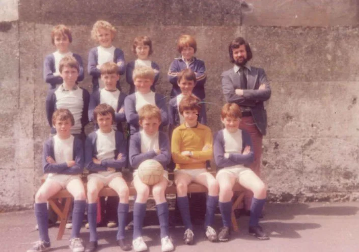 A vintage school photo of a boys' soccer team and their coach, with children wearing soccer uniforms and a coach in a blazer, in front of a concrete wall.