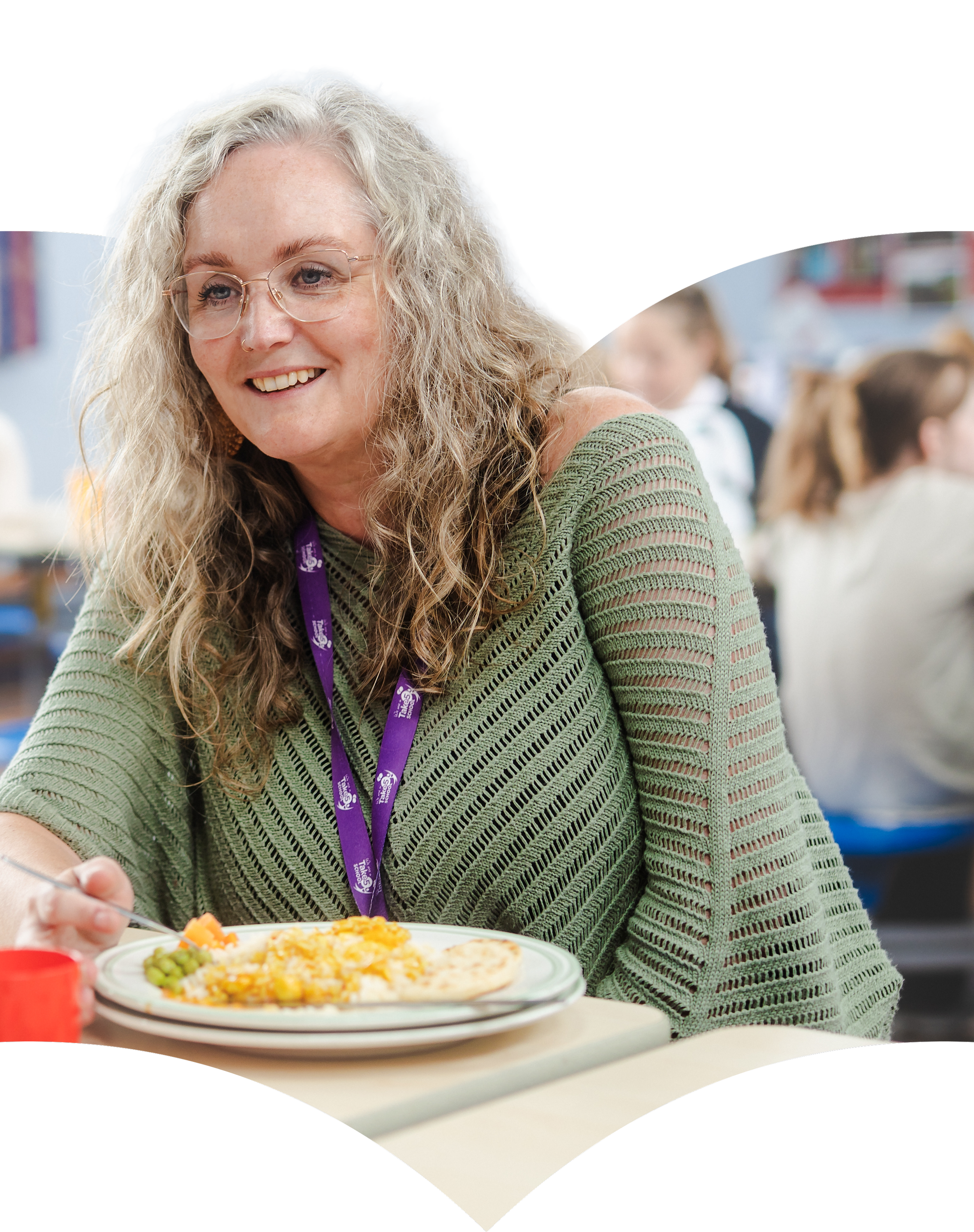 A woman with curly blonde hair, glasses, and a striped green sweater is smiling while serving herself food from a plate. The background shows a group of people in a social setting, possibly a cafeteria or dining hall.