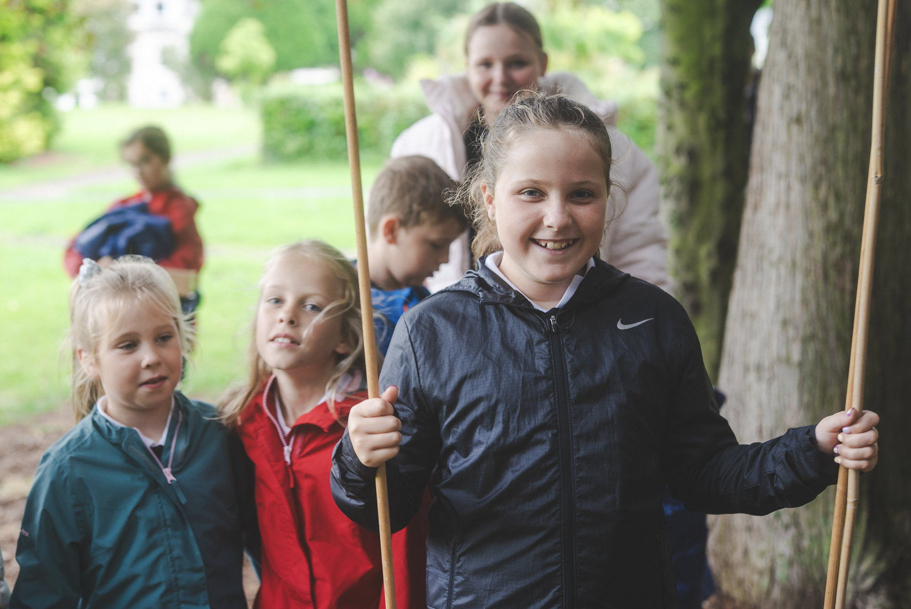 A group of children with an adult woman outdoors, posing for a photo, holding wooden sticks, near trees and greenery.