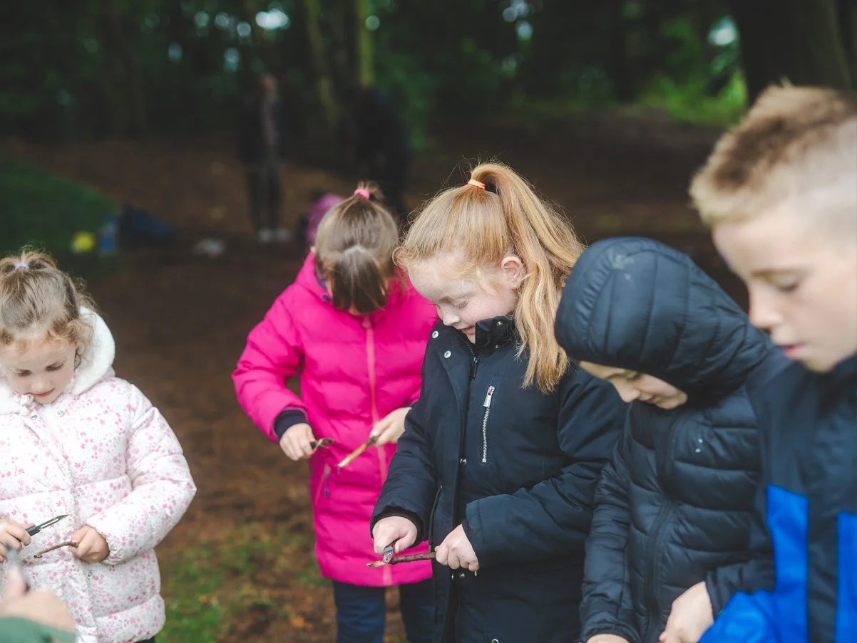 Group of children outdoors in a forest, wearing jackets, holding sticks, and looking down.