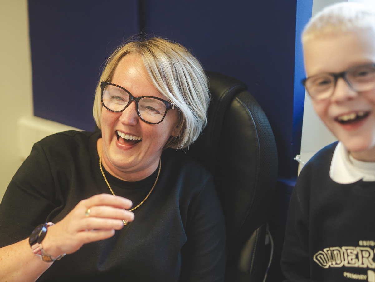 A woman with short blonde hair and glasses laughing, sitting in a chair, with a young boy with glasses and blonde hair next to her also smiling.