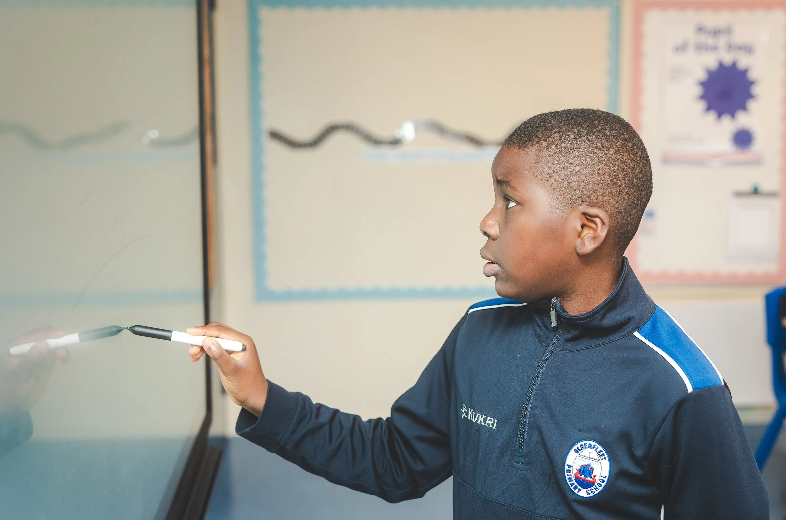 A young student in a school uniform writing on a board with a marker in a classroom.